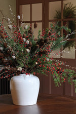 Decorative arrangement of greenery, red berries, and pinecones in a white vase on a wooden surface.