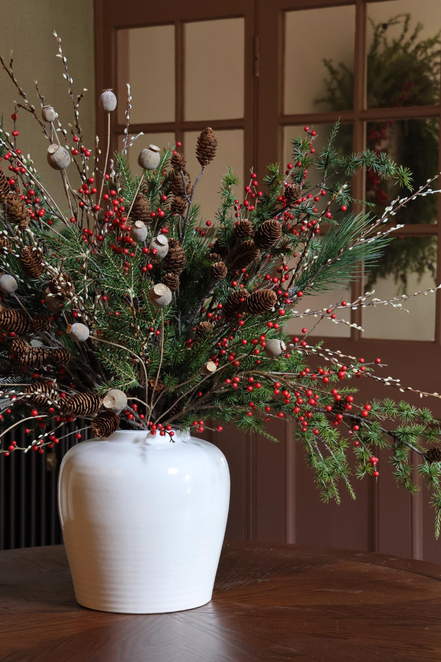 Decorative arrangement of greenery, red berries, and pinecones in a white vase on a wooden surface.
