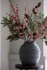 Decorative arrangement of greenery, red berries, and pine cones in a textured grey vase on a wooden stool.