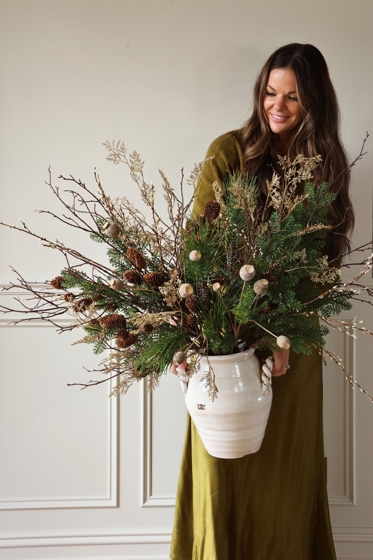 women in dark green dress holding a white ceramic vase with artficiail green foliage and twigs 