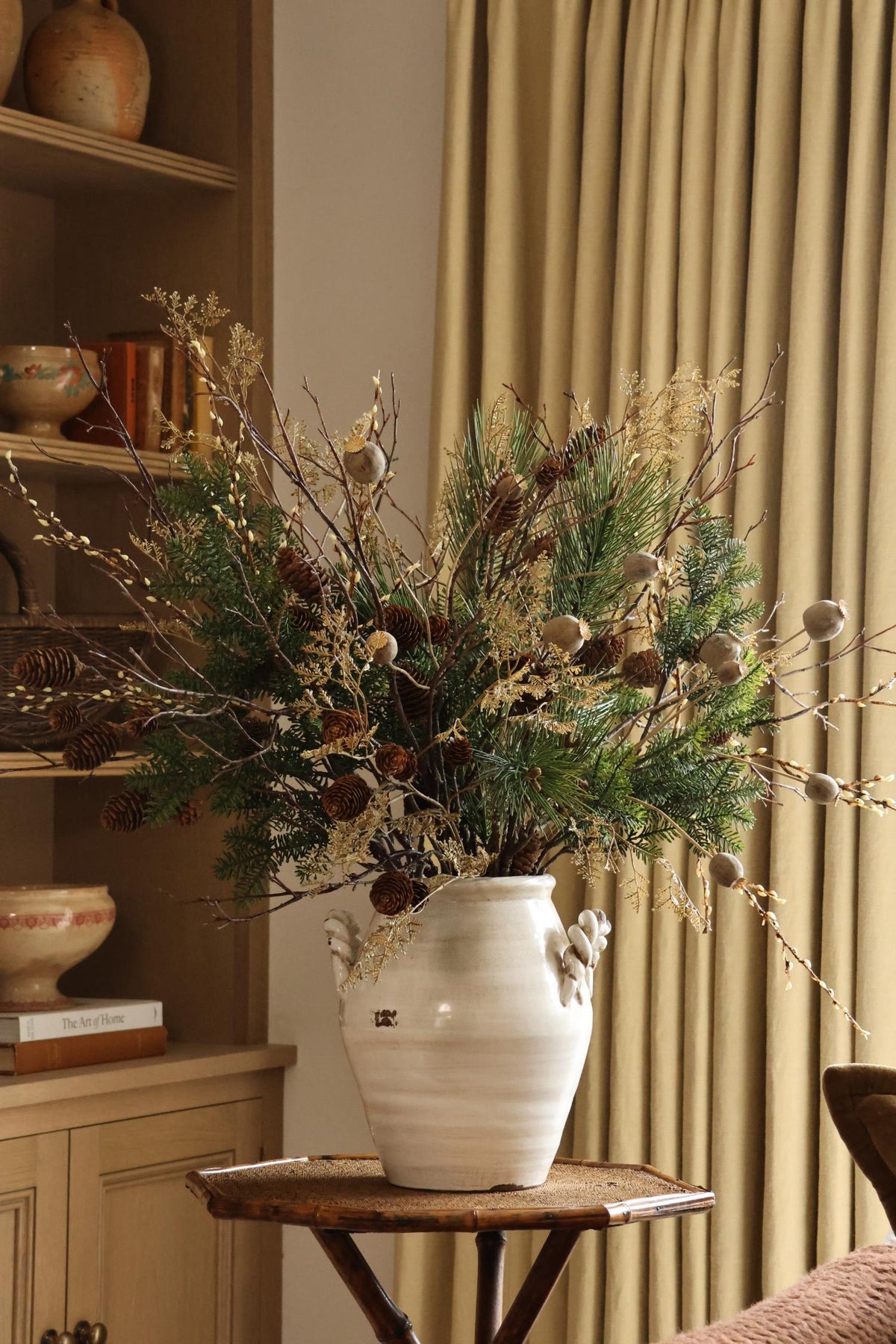 Large white ceramic vase with artificial winter foliage arrangement on small table with curtains and wood shelving behind 