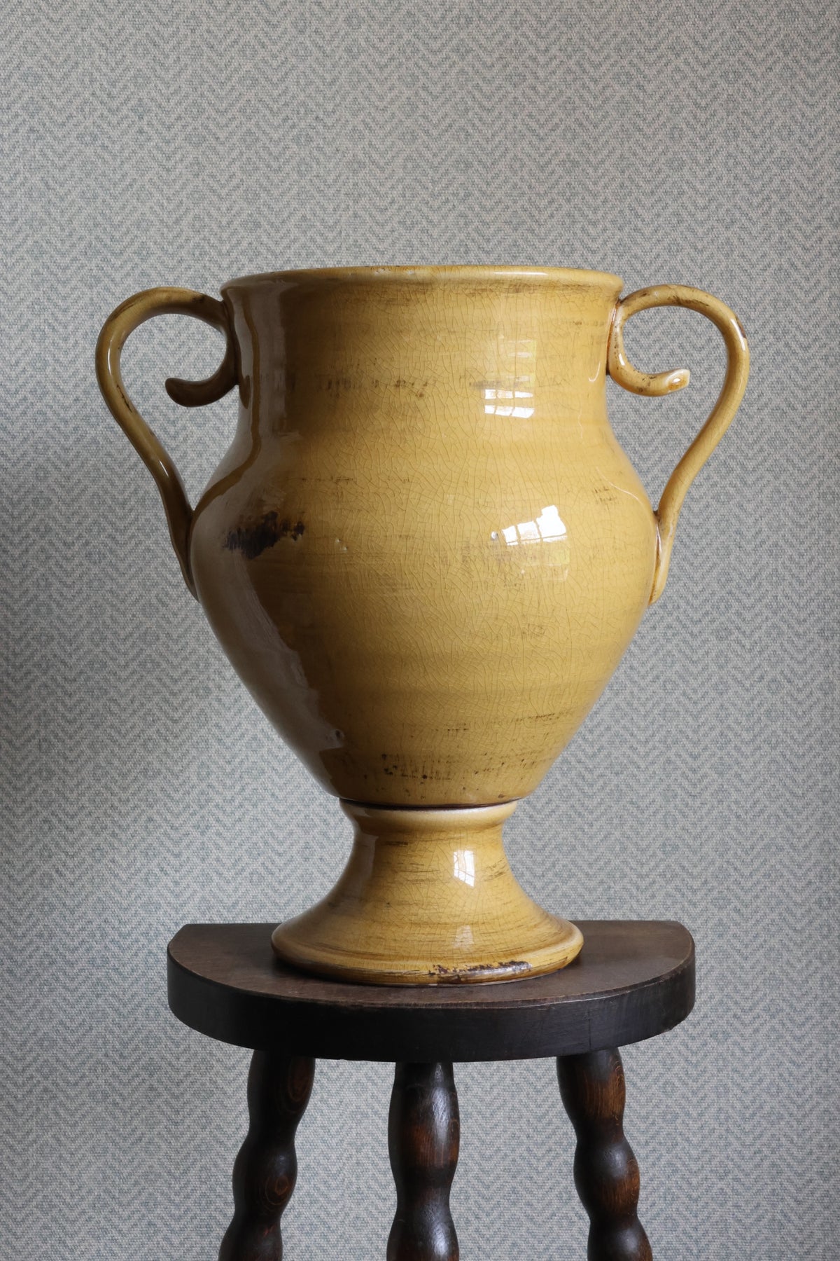 Yellow ceramic urn with handles on a wooden stand against a gray textured wall.