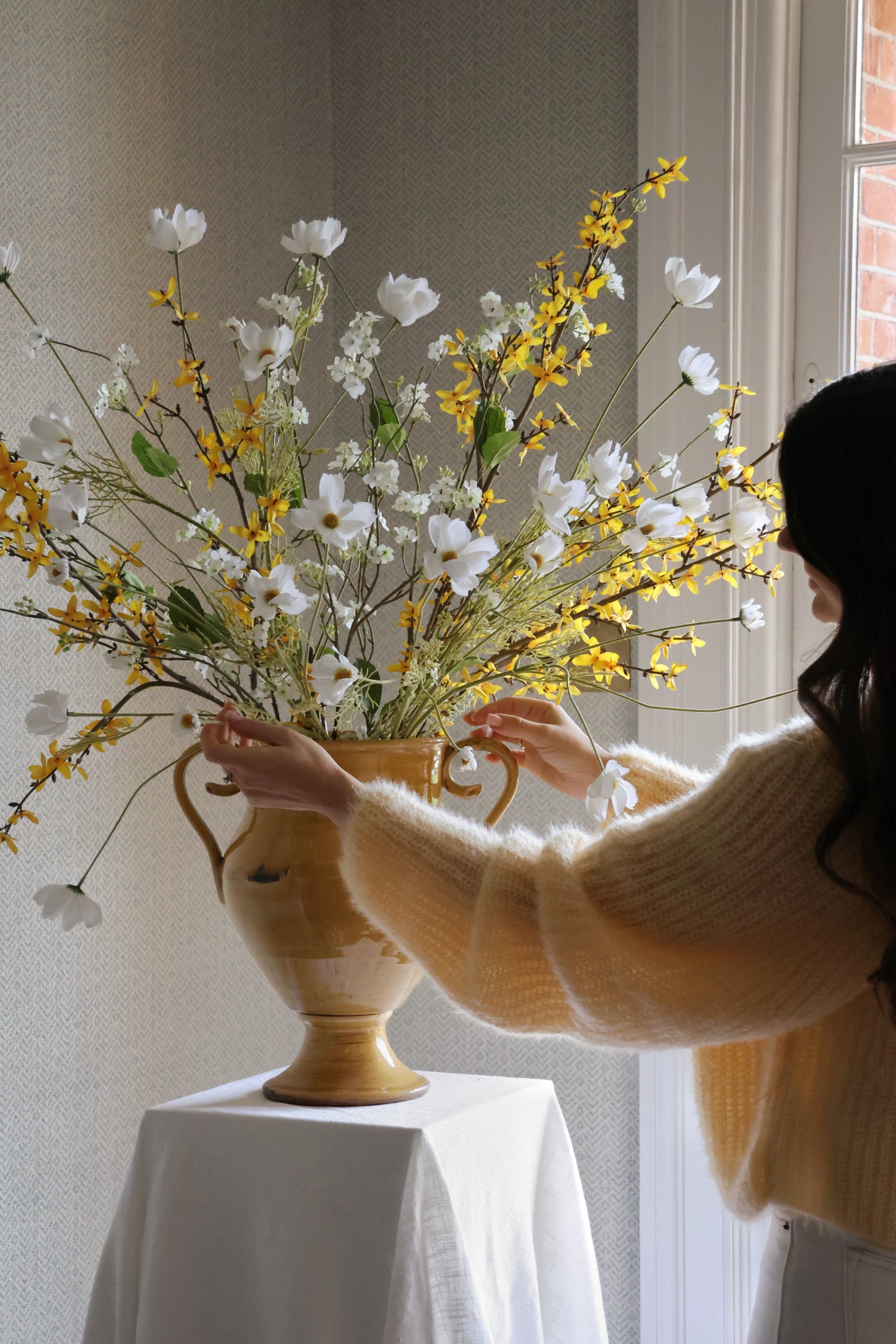 Lady with yellow cardigan arranging flowers in a yellow urn with white and yellow flowers