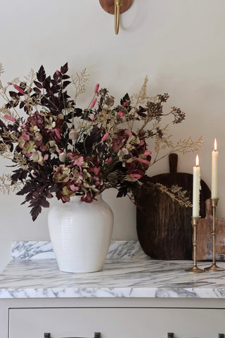 White vase with dried flowers on a marble surface with candles and a wooden board in the background.