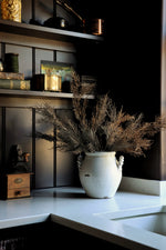 A rustic white ceramic vase filled with dried brown grasses sits on a white countertop in a dark kitchen setting with shelves and cabinets behind it.