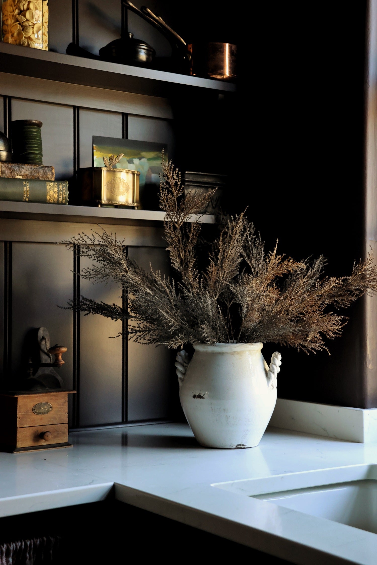 A rustic white ceramic vase filled with dried brown grasses sits on a white countertop in a dark kitchen setting with shelves and cabinets behind it.