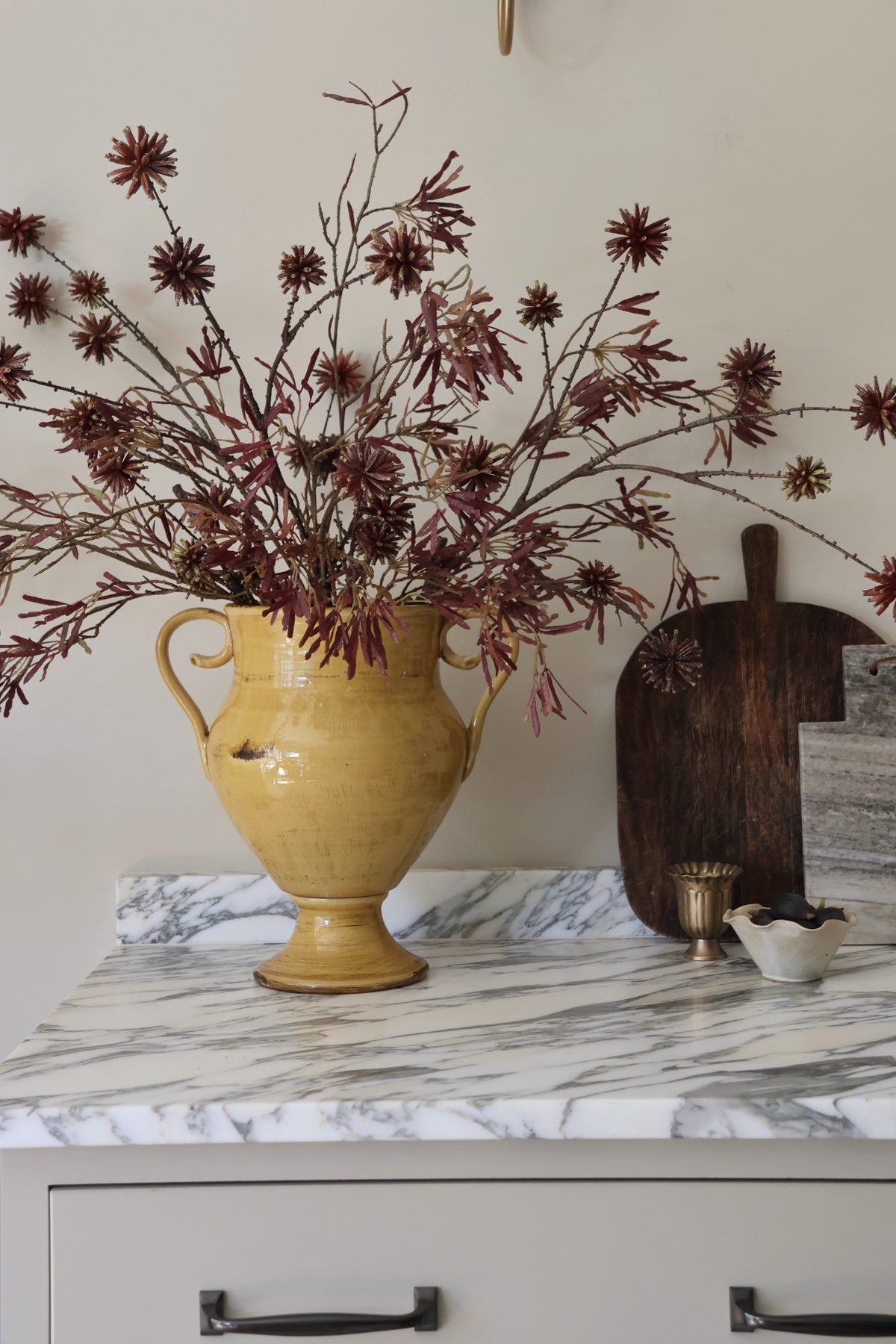 A yellow ceramic vase filled with dried burgundy-brown pinecones and branches, styled on a white marble countertop next to a dark wooden cutting board and a small white dish.