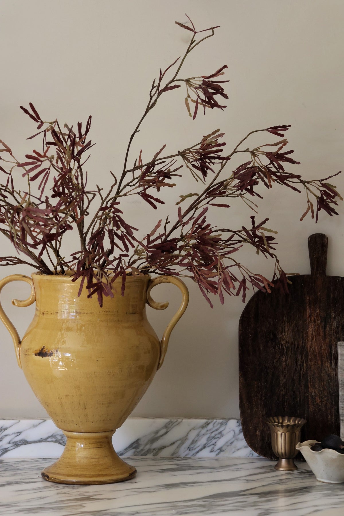 A rustic yellow ceramic vase with two handles, filled with dried burgundy foliage, set on a white marble surface next to a dark wooden cutting board and a small brass cup.