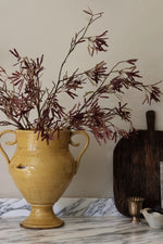 A rustic yellow ceramic vase with two handles, filled with dried burgundy foliage, set on a white marble surface next to a dark wooden cutting board and a small brass cup.