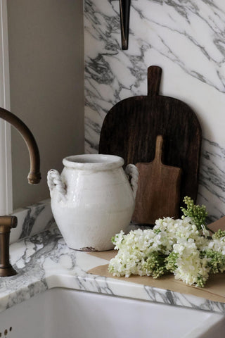 White ceramic vase on a kitchen counter with marble backsplash and cutting board.