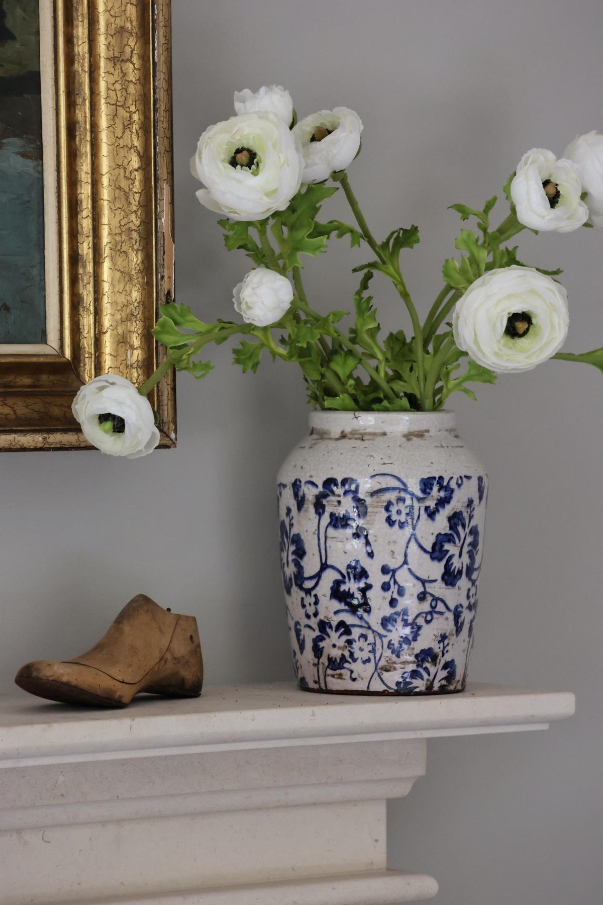 White flowers in a blue and white vase on a mantelpiece with a framed picture in the background.