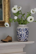 White flowers in a blue and white vase on a mantelpiece with a framed picture in the background.