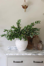 Decorative setup with a white vase containing faux olive stems on a marble surface.