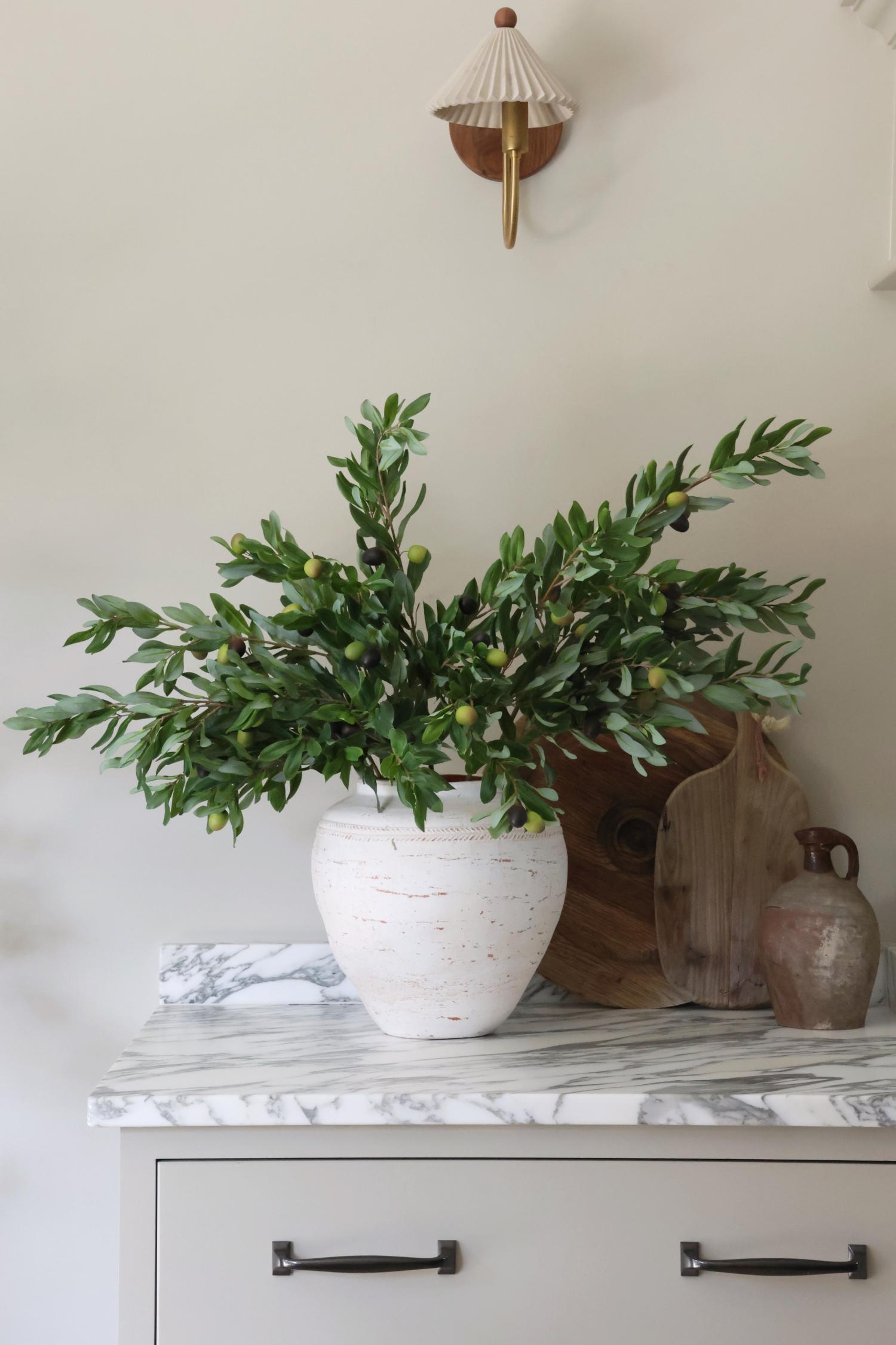 Decorative setup with a white vase containing faux olive stems on a marble surface.