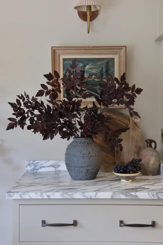 Decorative setting with a vase, plant, and fruit on a marble surface with a painting in the background.
