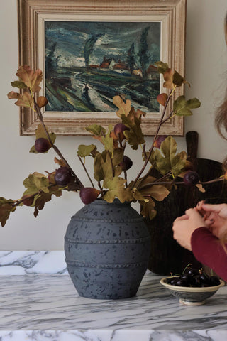 Decorative vase with branches and figs on a marble surface, framed painting in the background