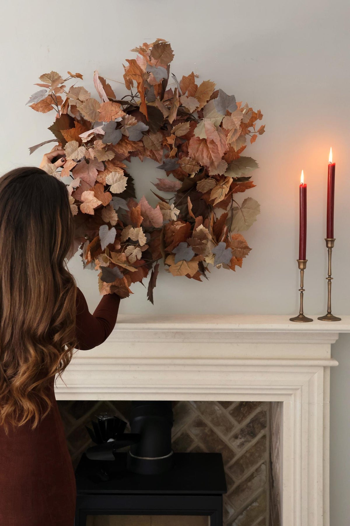 Person adjusting a decorative autumn wreath on a fireplace mantel with candles.