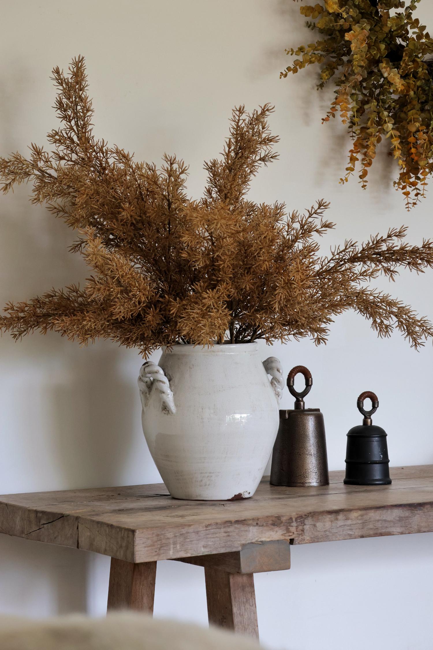 White vase with faux autumn foliage on a wooden table against a neutral wall styled with bells
