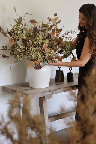 Woman arranging artificial flowers in a white vase on a wooden table.
