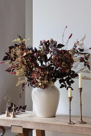 Decorative floral arrangement in a white vase on a wooden table with candles and small decorative items.