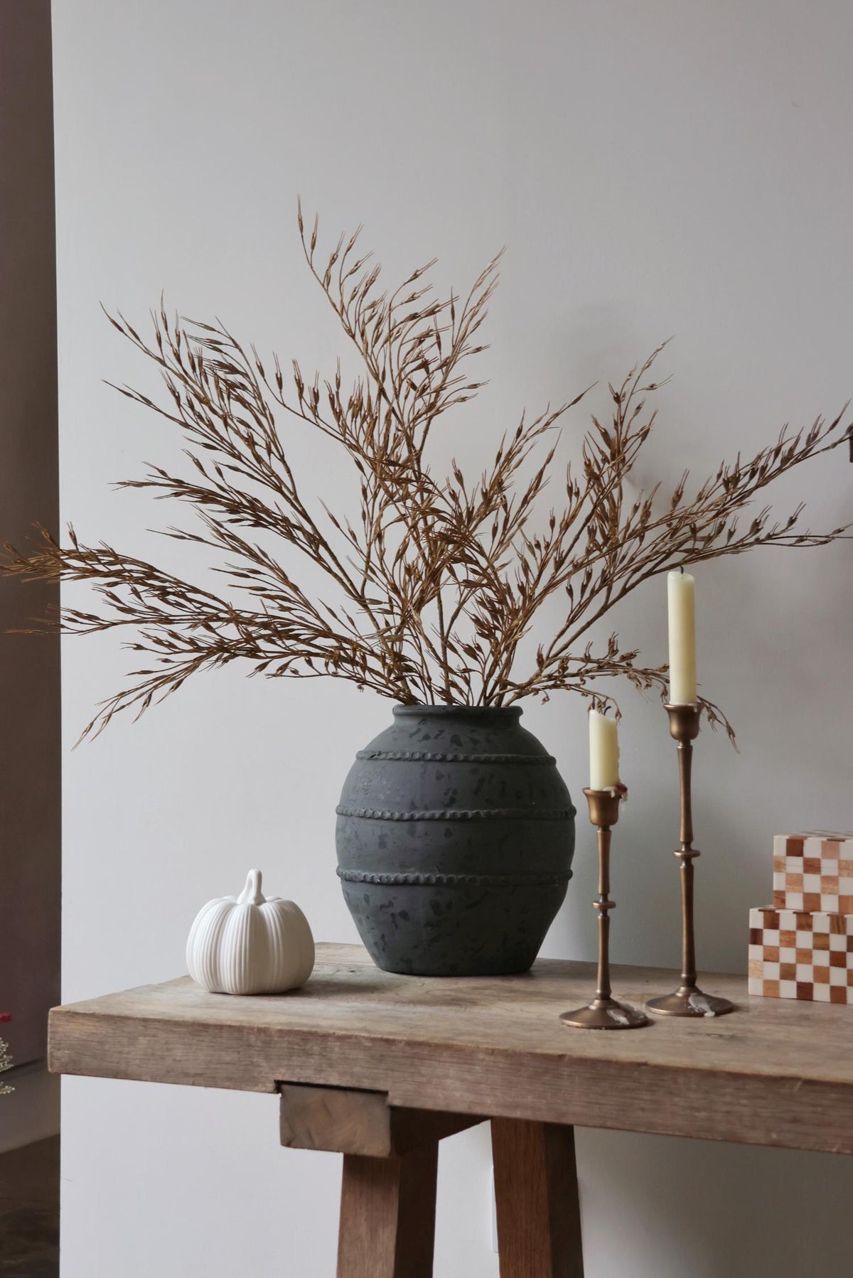 Decorative setup with a dark textured vase, faux wheat grass, white pumpkin, and candles on a wooden table against a plain wall.