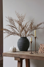 Decorative setup with a dark textured vase, faux wheat grass, white pumpkin, and candles on a wooden table against a plain wall.