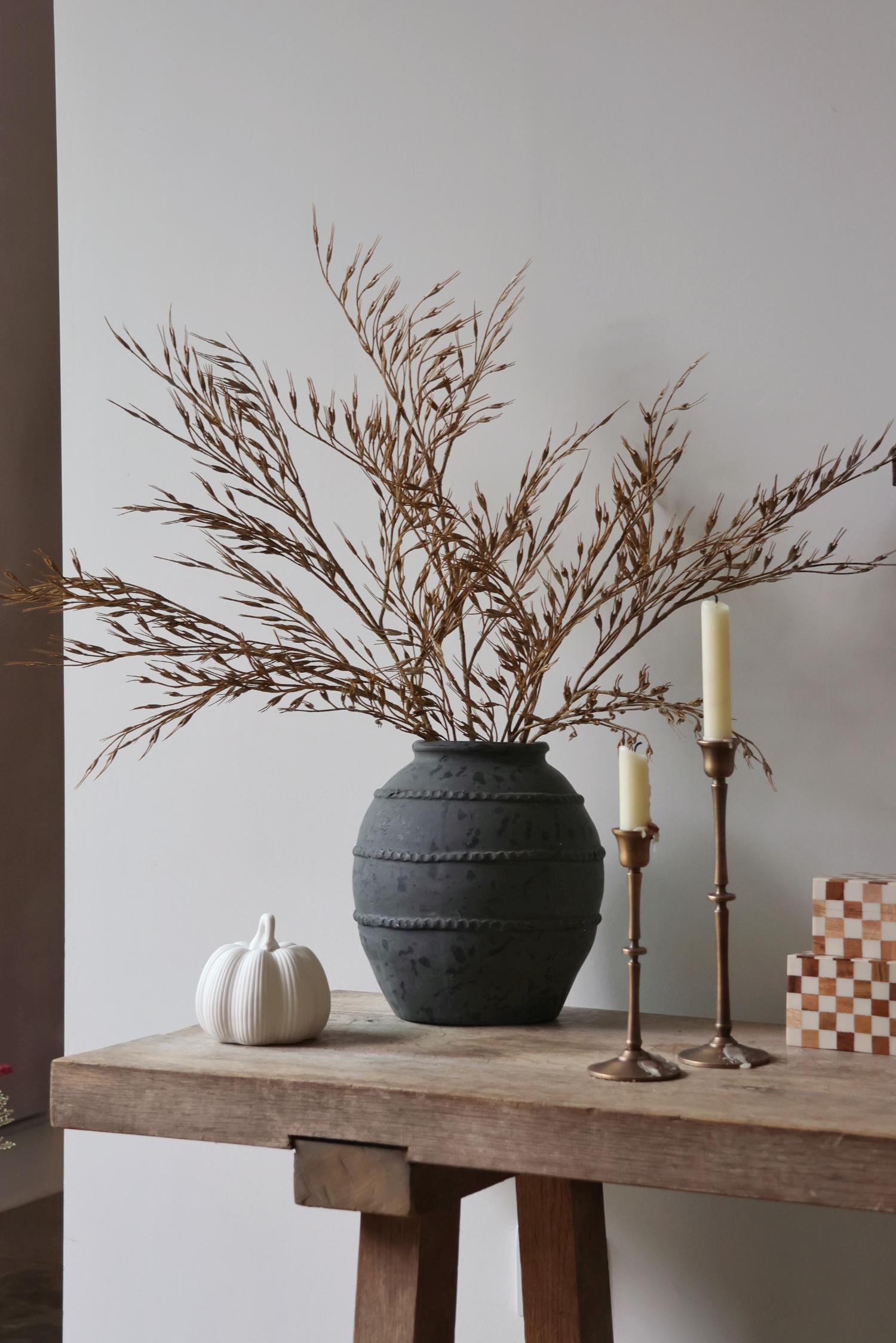 Decorative setup with a dark textured vase, faux wheat grass, white pumpkin, and candles on a wooden table against a plain wall.
