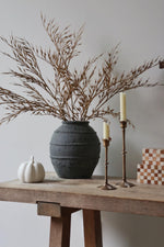 Decorative setup with a textured black vase, dried branches, candles, and a small pumpkin on a wooden surface.
