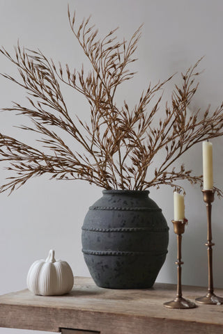 Decorative setup with a dark textured vase, faux wheat grass, white pumpkin, and candles on a wooden table against a plain wall.