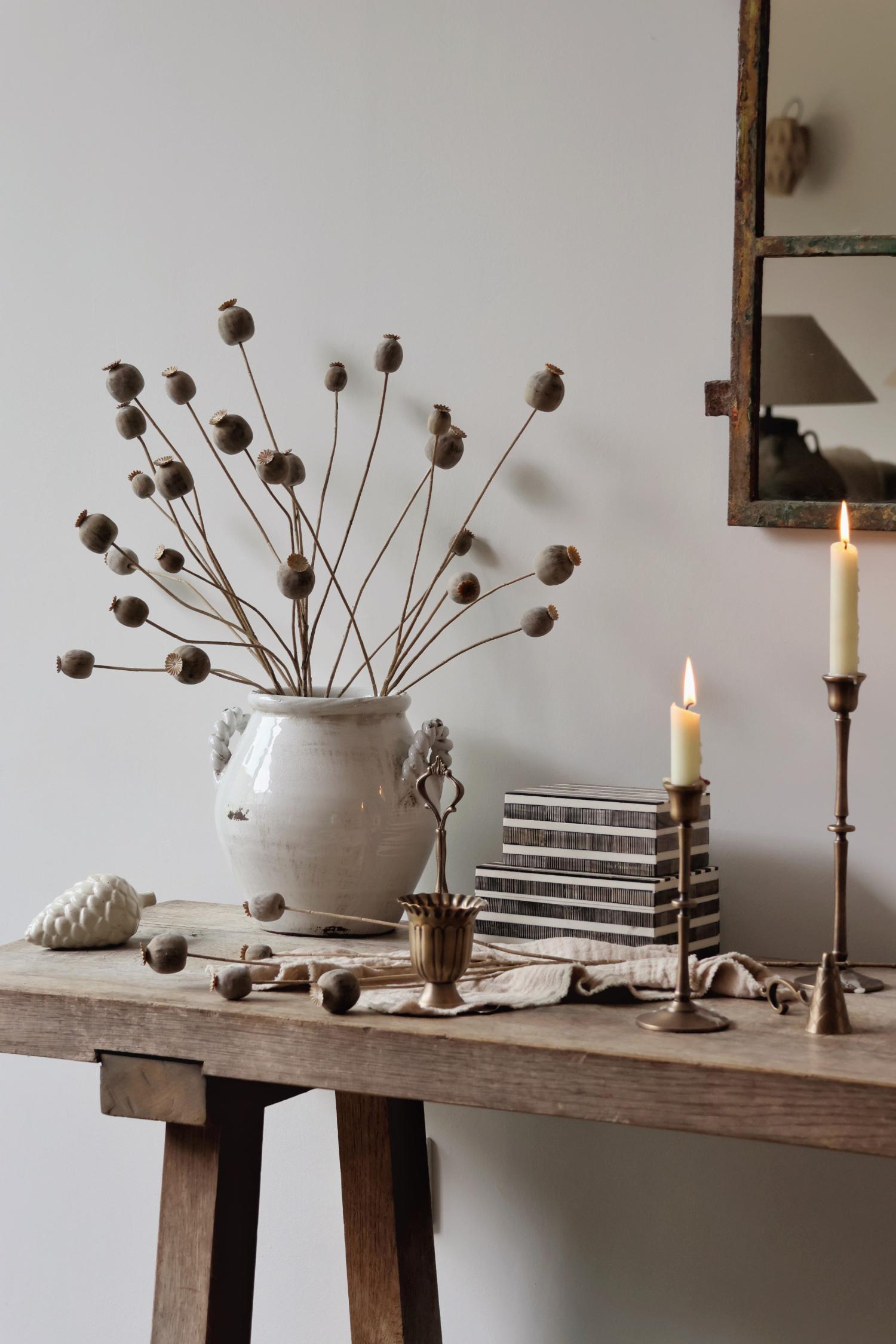 Decorative setting with a vase, candles, and faux dried poppy heads on a wooden table.