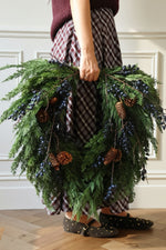 Women standing to the side holding an artificial wreath with dark blue berries, pine cones and branches 