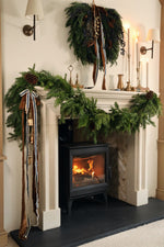 large artificial juniper wreath with blue berries, pinecones and branches with hanging brown and blue ribbon and a cluster of small hanging brass bells. Positioned on a cosy living room mantle with a mixed foliage garland draping over one side
