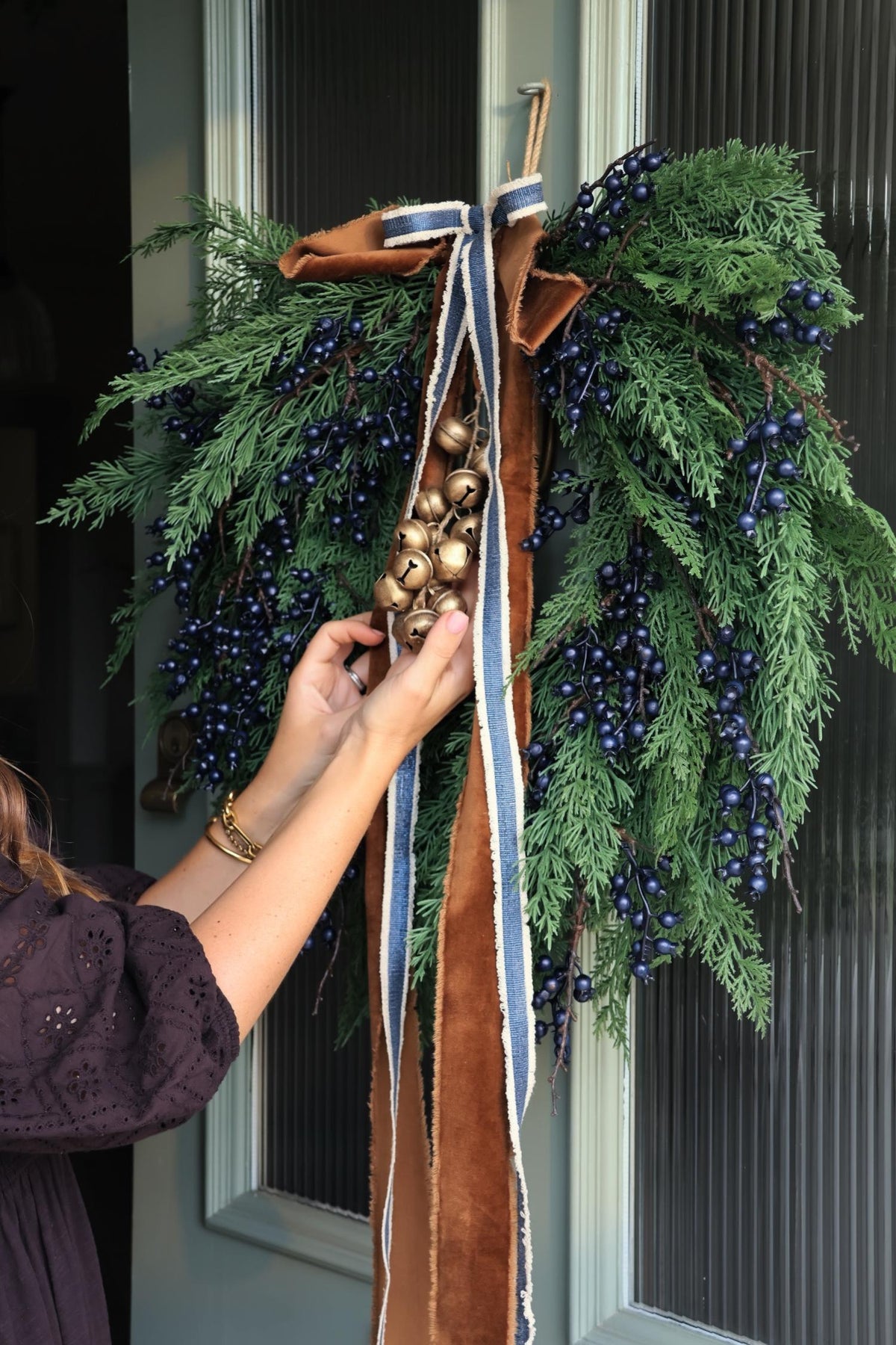 large artificial wreath with blueberries on a door with brown and blue velvet ribbons and brass bells.