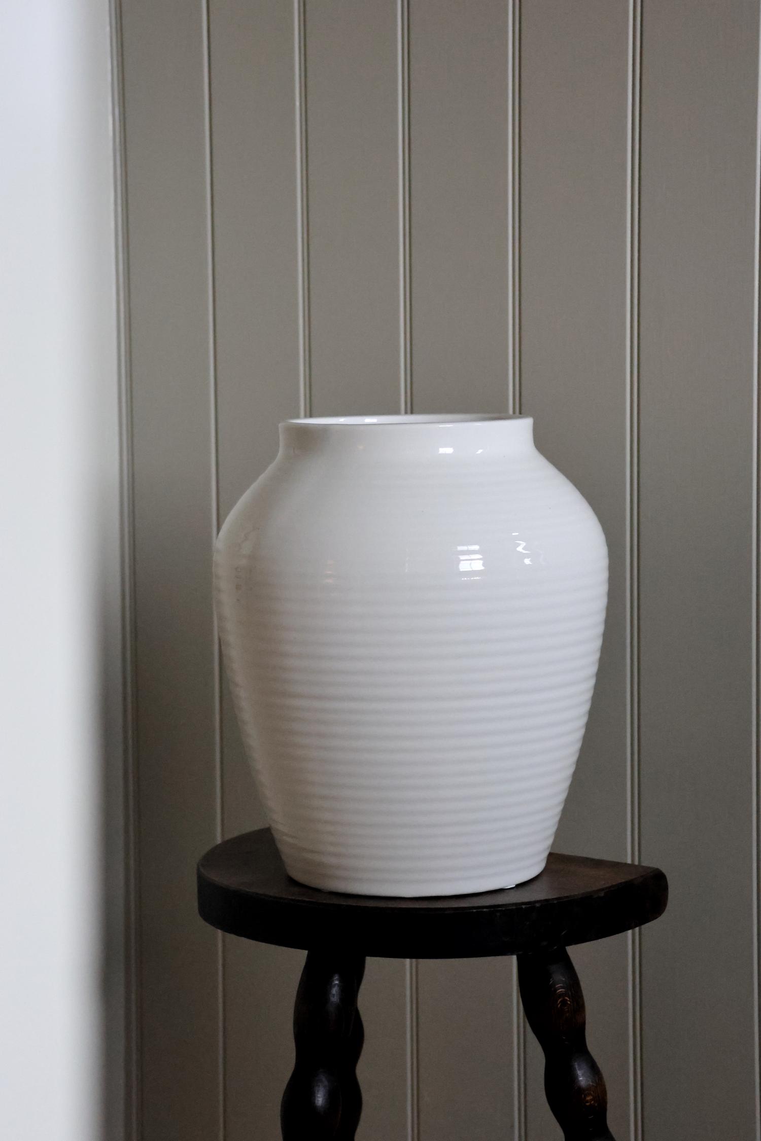 A white, ribbed, tapered ceramic vase placed on a dark wooden stool against a wall with vertical panels.