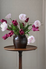 Bouquet of pink and purple tulips in a dark vase on a wooden table against a neutral wall.