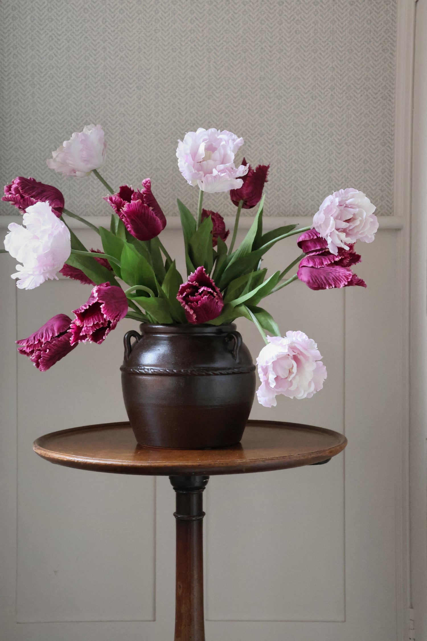 Bouquet of pink and purple tulips in a dark vase on a wooden table against a neutral wall.