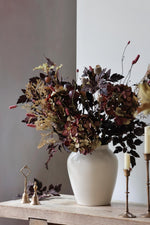 White vase with autumn artificial flower arrangement in deep burgundy colour tones styled with brass bells and candlesticks on a wooden surface 