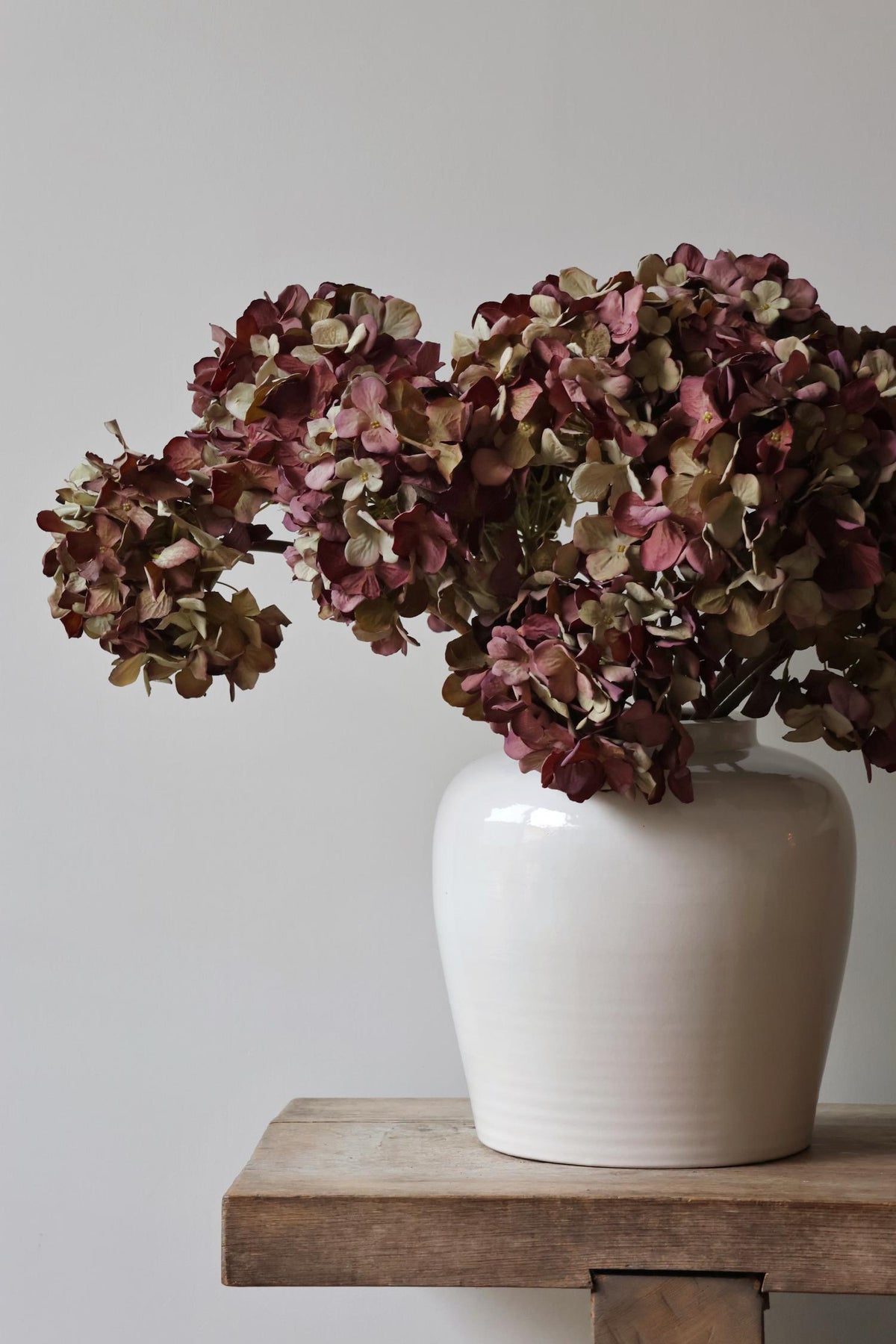 White vase with artificial dried burgundy hydrangeas on a wooden surface against a plain background