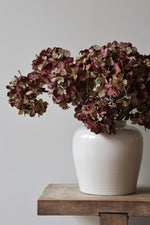 White vase with artificial dried burgundy hydrangeas on a wooden surface against a plain background