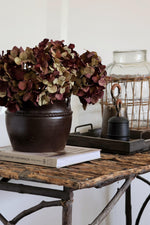 Decorative setup with a vase of deep burgundy artificial dried look hydrangea, books, and a lantern on a rustic table.