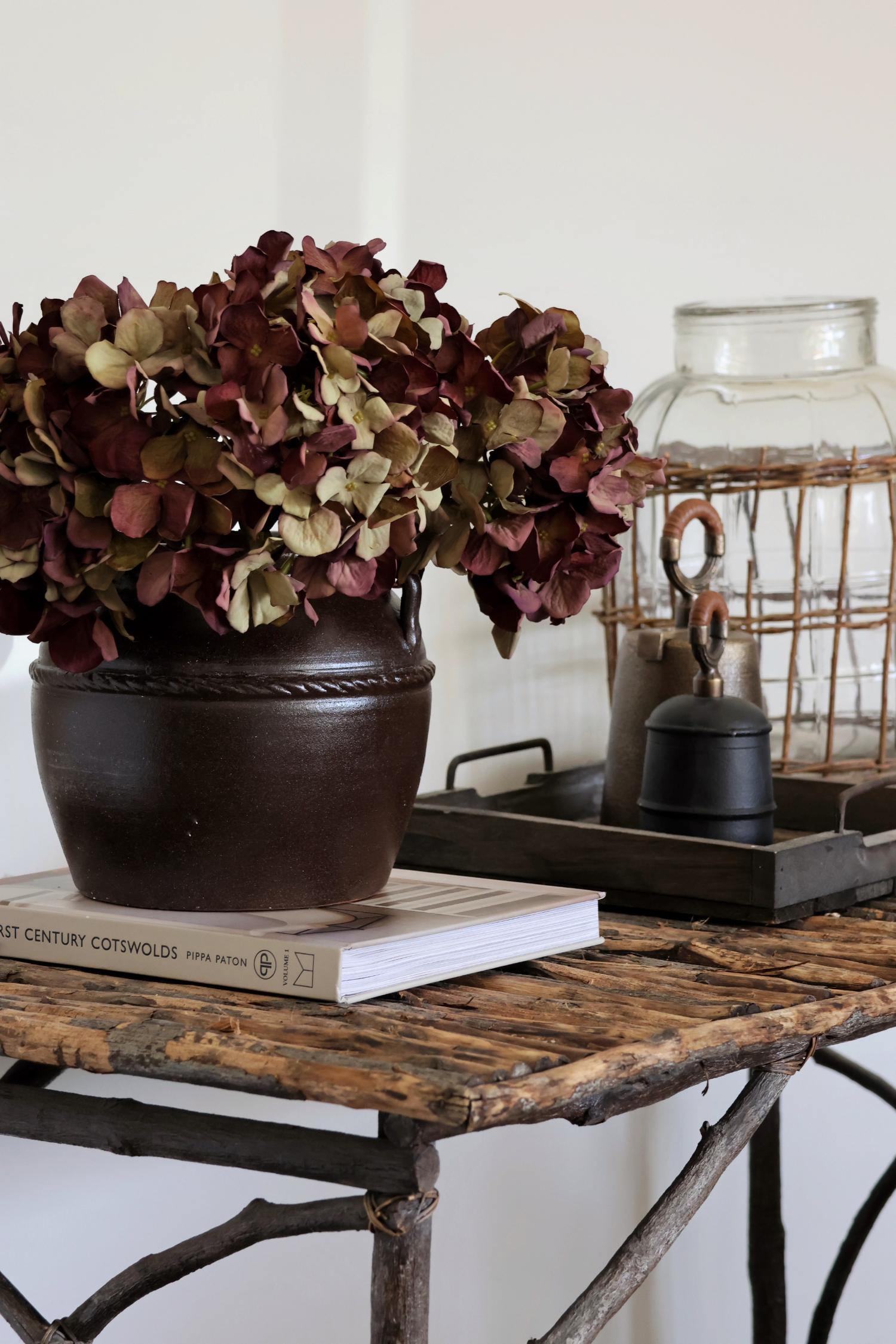 Decorative setup with a vase of deep burgundy artificial dried look hydrangea, books, and a lantern on a rustic table.