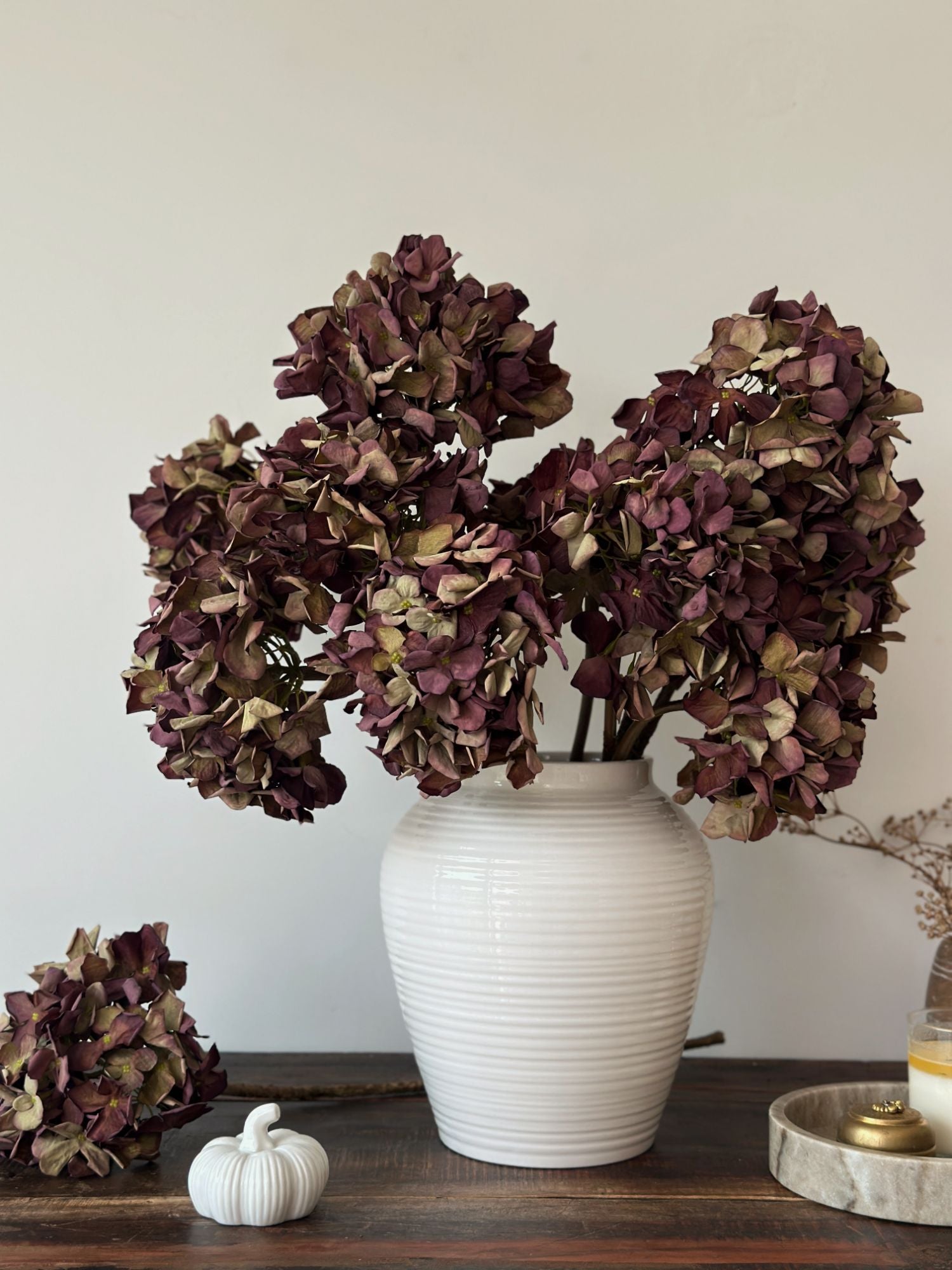white ceramic vase with artificial faux dried burgundy hydrangeas on a wooden surface with white pumpkin