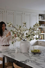 A faux pear blossom arrangement displayed in a white vase on a marble countertop, with a person standing in the background.