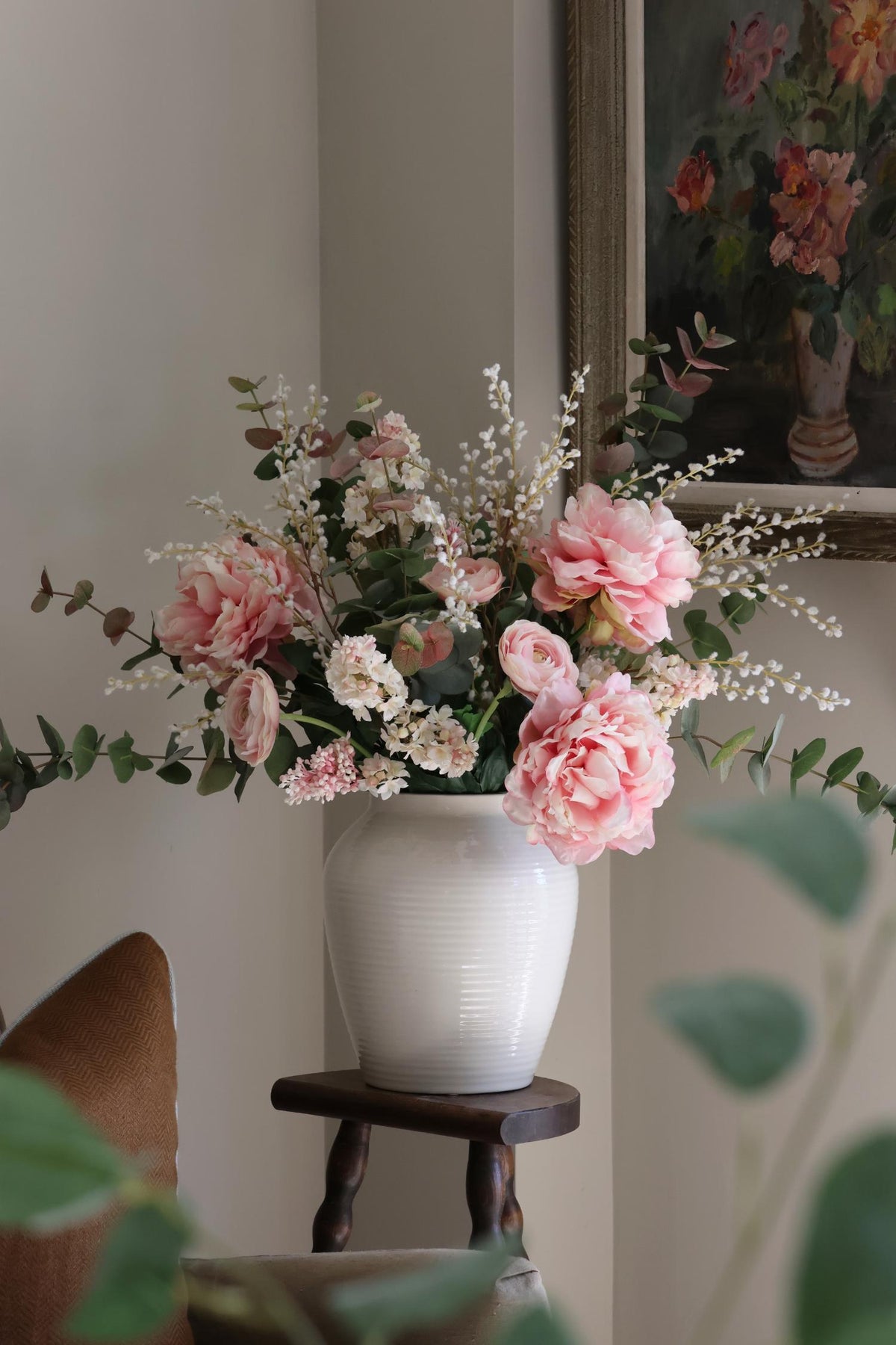 A faux pink peony arrangement in a white vase, placed on a wooden stool in a room with a floral wallpaper.