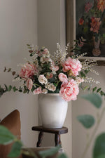 A faux pink peony arrangement in a white vase, placed on a wooden stool in a room with a floral wallpaper.