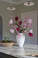 White vase with pink and purple flowers on a marble countertop with two hanging light fixtures.
