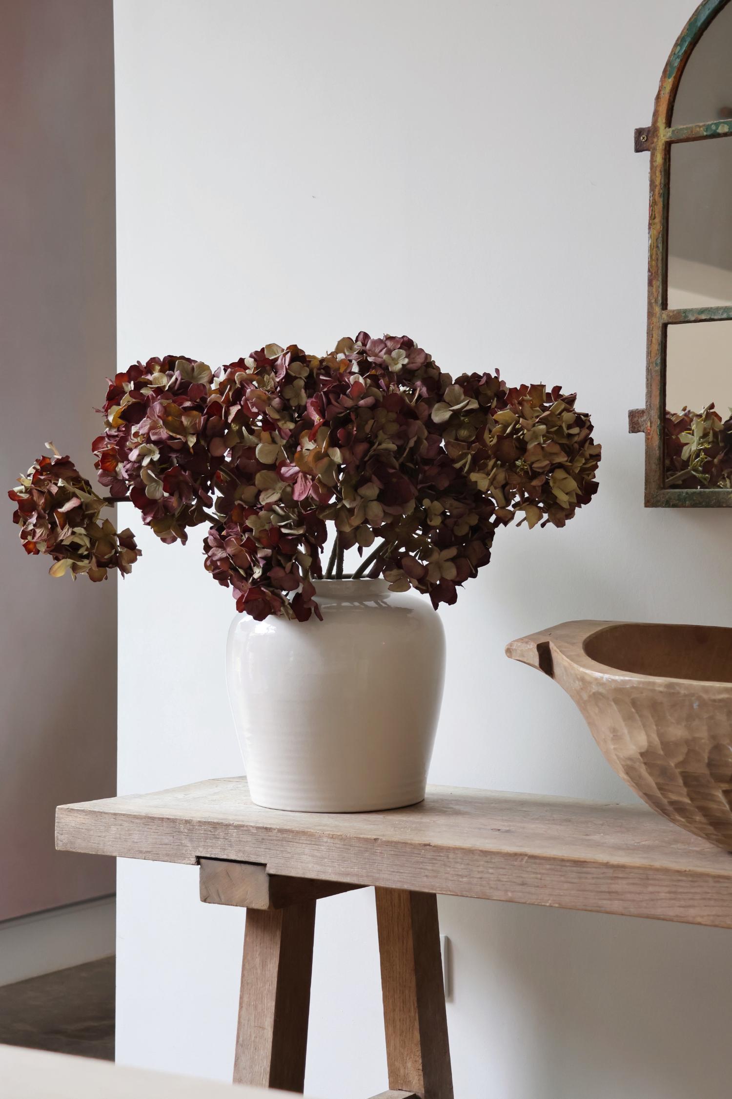 White suffolk vase with faux mulberry dried hydrangea flowers on a wooden table against a white wall.