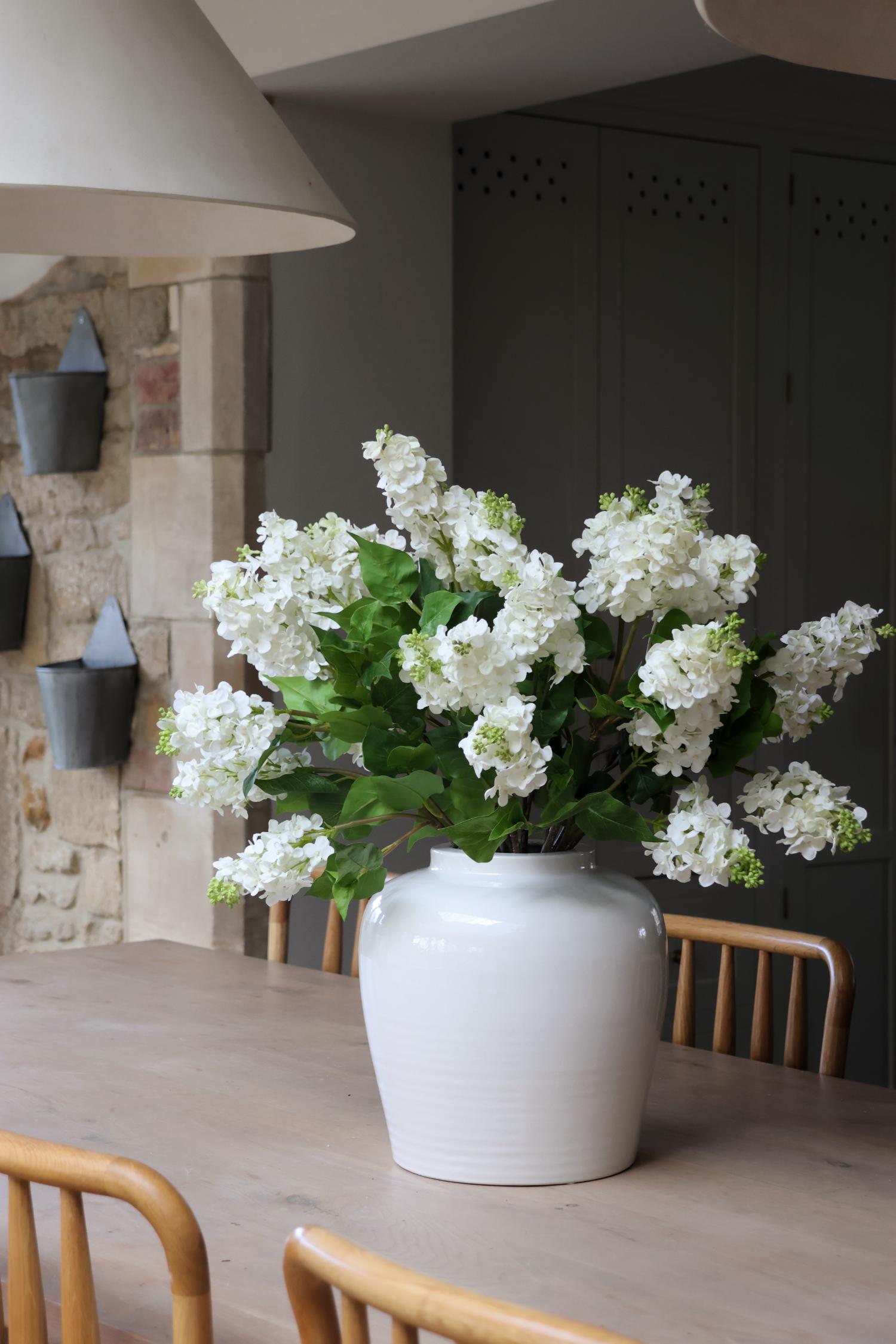 large white ceramic Suffolk Vase and faux white lilacs with large green leaves on a wooden dining table 