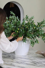 Person arranging artificial green olive branches in a white ceramic vase on a marble surface