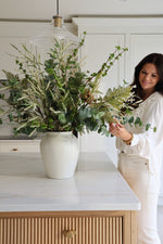 Woman arranging flowers in a kitchen with a white vase and greenery.